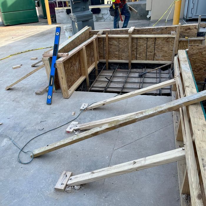 Construction site with an open rectangular pit, wooden formwork, and metal supports on a concrete floor.