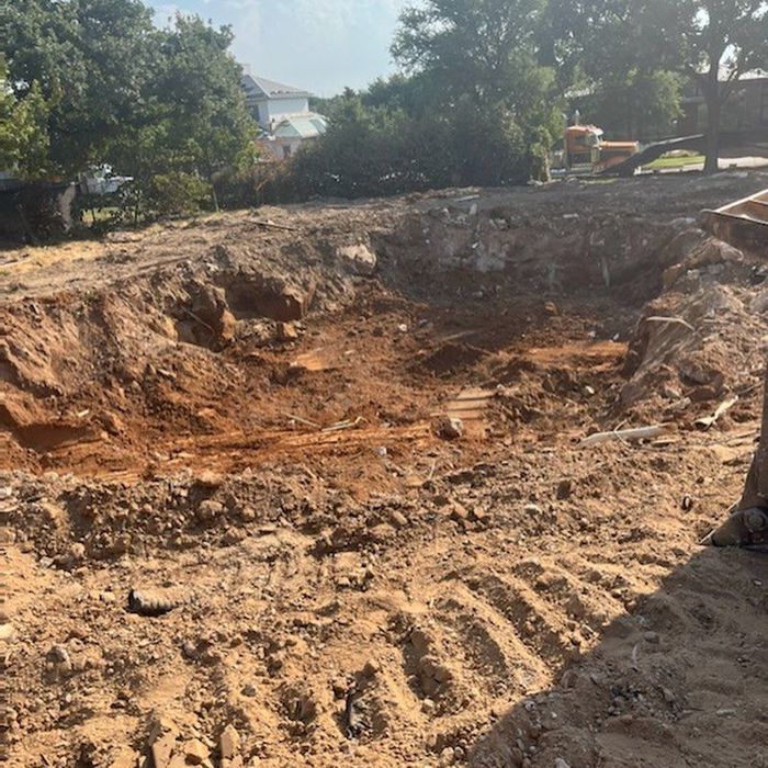 Excavated dirt pit with exposed soil and tire tracks, surrounded by trees and houses in the background