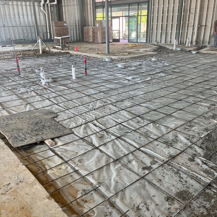 Rebar grid over plastic sheeting in an unfinished indoor construction floor, with plumbing stubs visible.