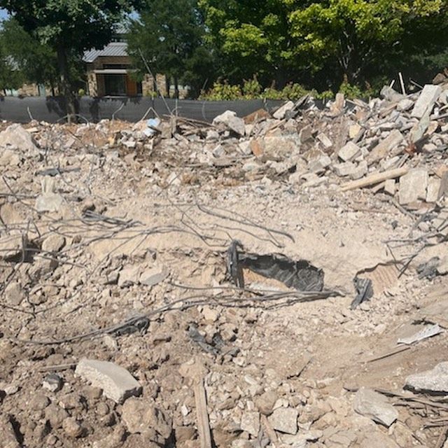 Rubble and broken concrete piled in a cleared lot with trees and a house in the background