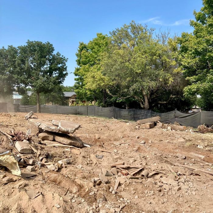 Cleared dirt lot with scattered rubble and a line of trees behind a temporary fence under a blue sky