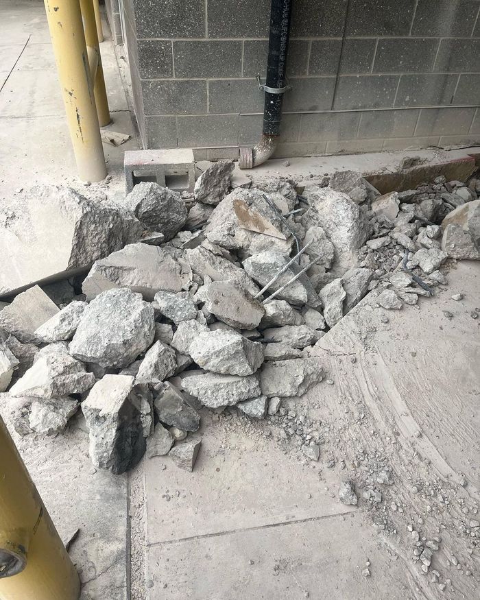 Pile of broken concrete rubble on a sidewalk beside a cinder block wall and yellow post.