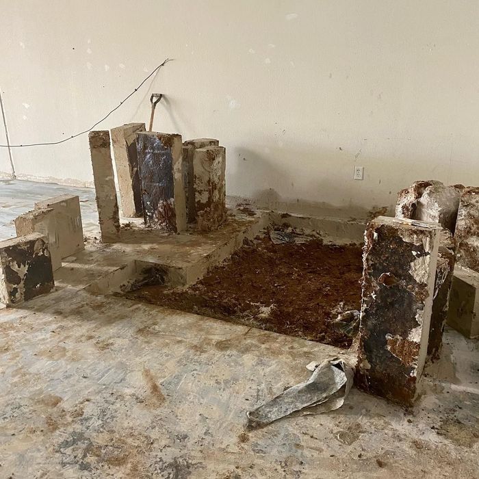 Damaged concrete basement with crumbling pillars, debris, and exposed soil patch on the floor