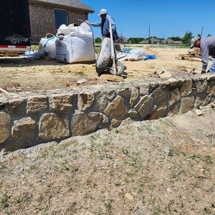 Workers building a stone retaining wall beside a house, with sandbags and construction materials nearby.