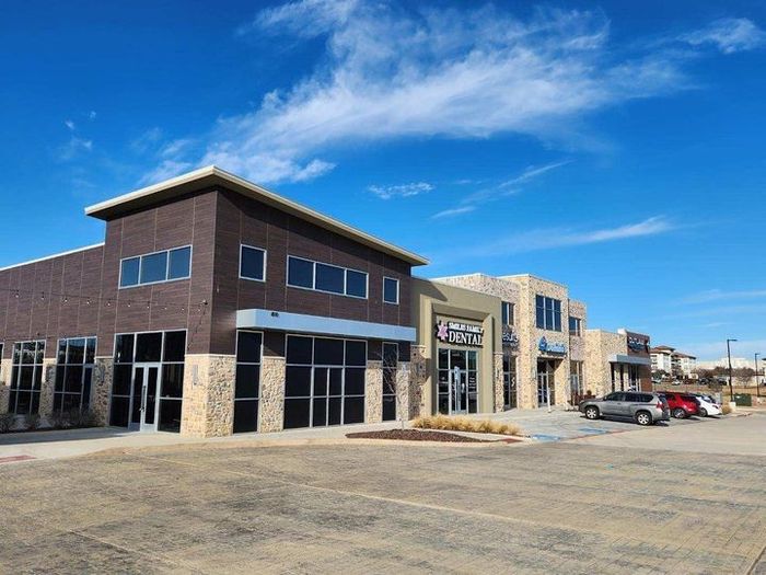 Strip mall storefronts under a bright blue sky, with a paved parking lot and a few parked cars.