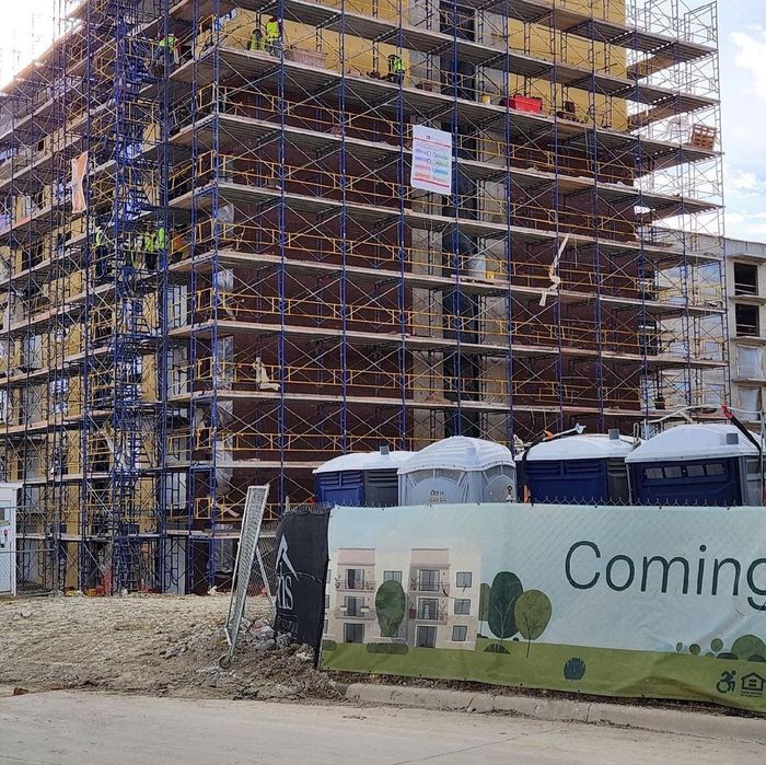 Scaffolded building under construction with portable toilets and a “Coming” site banner in front.