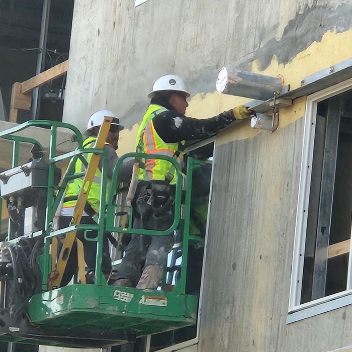 Two workers in a green lift apply material to a concrete wall at a construction site