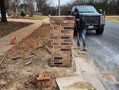 Worker repairing a brick mailbox beside a driveway with a black truck nearby