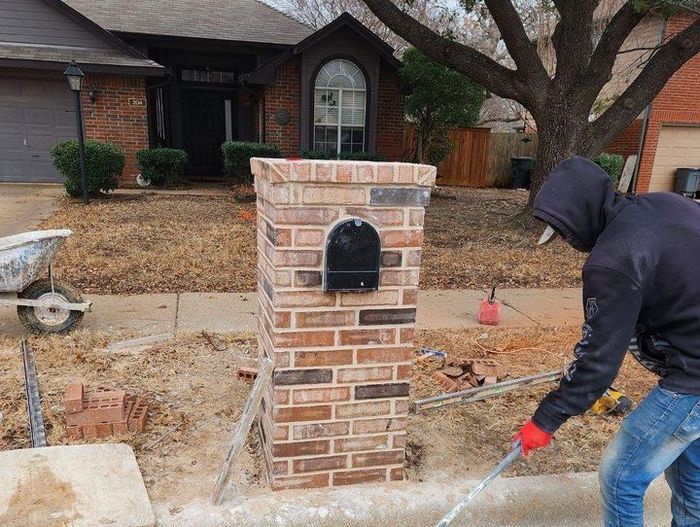 Person in a hooded jacket cleaning a brick mailbox with a pressure washer in a suburban yard