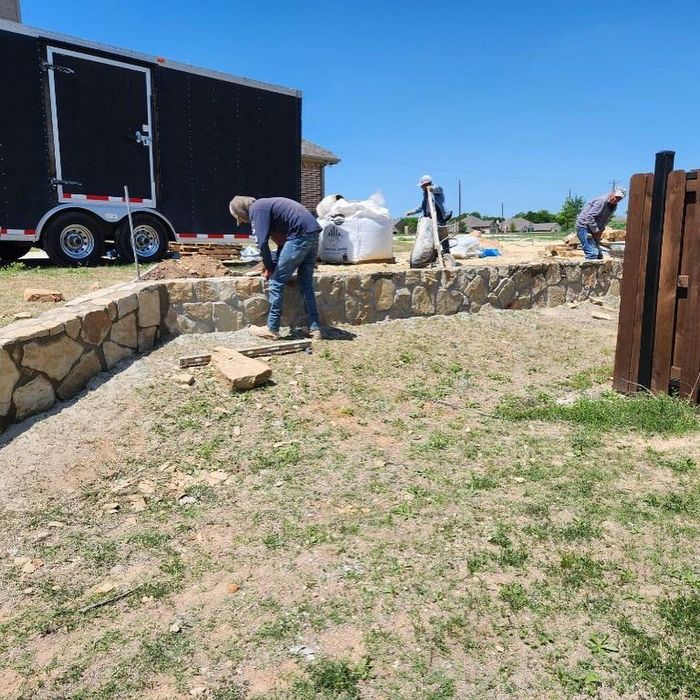 Workers building a stone retaining wall beside a black trailer on a sunny day