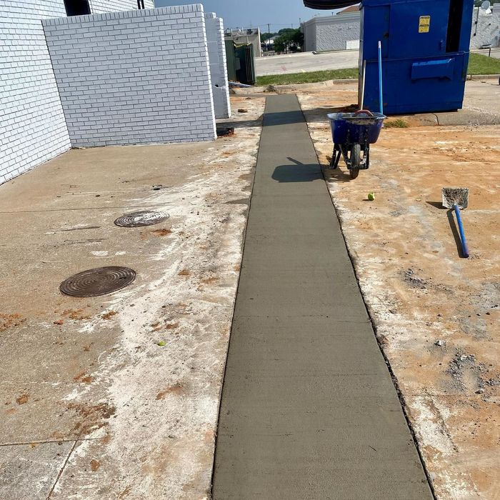 Freshly poured concrete walkway beside white wall and blue building, with wheelbarrow at the end.