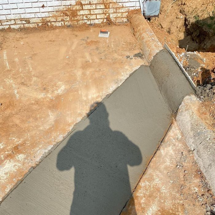 Fresh gray concrete walkway being poured beside a brick wall at a construction site.