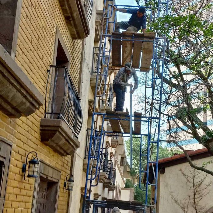 Two workers on a tall scaffold beside a yellow brick building, one standing on a platform under a tree.