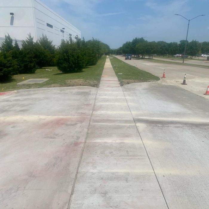 Empty concrete walkway beside a white building, lined with trees and traffic cones on a sunny day