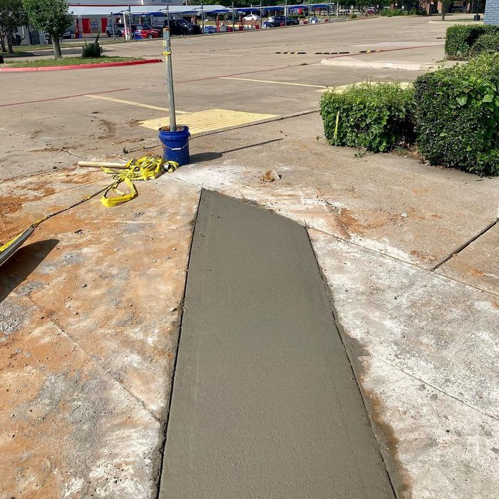Freshly poured concrete patch on a sidewalk near a parking lot, with a blue bucket and yellow tape nearby