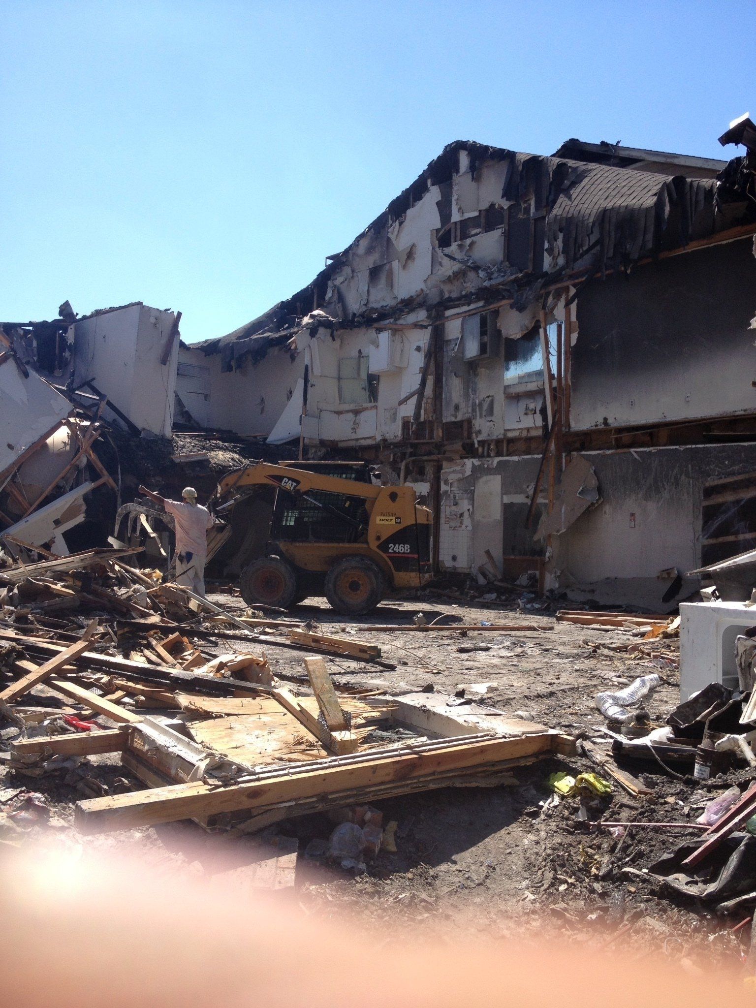 Collapsed building ruins with debris and a yellow construction loader under a clear blue sky