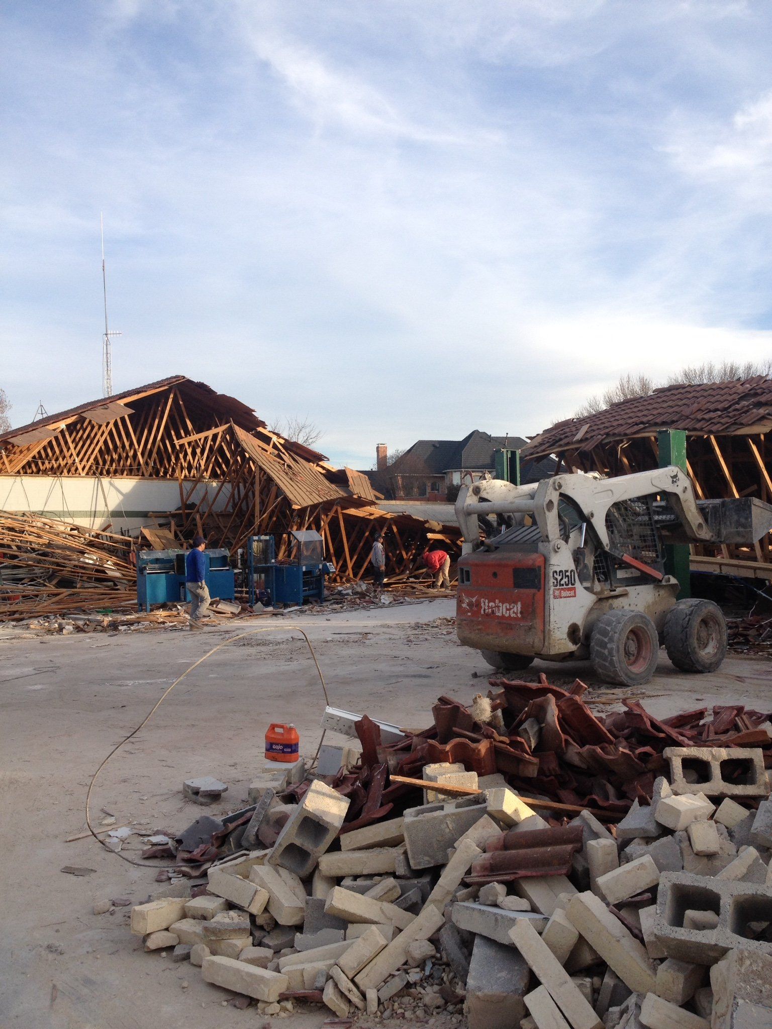 Storm-damaged building site with debris, a skid-steer loader, and exposed roof trusses under a cloudy sky
