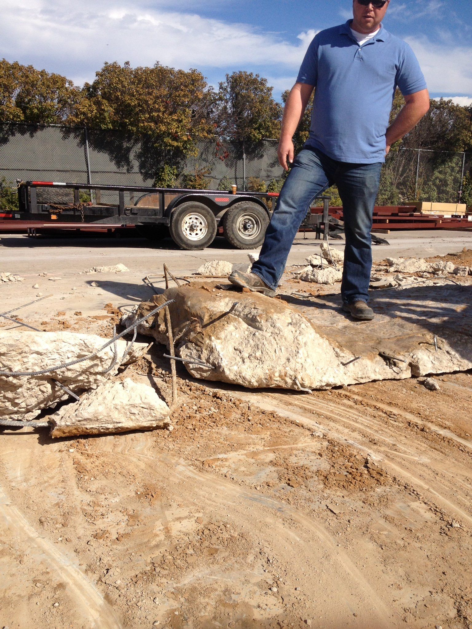 Man standing on large rocks at a dusty construction site beside a trailer and trees.