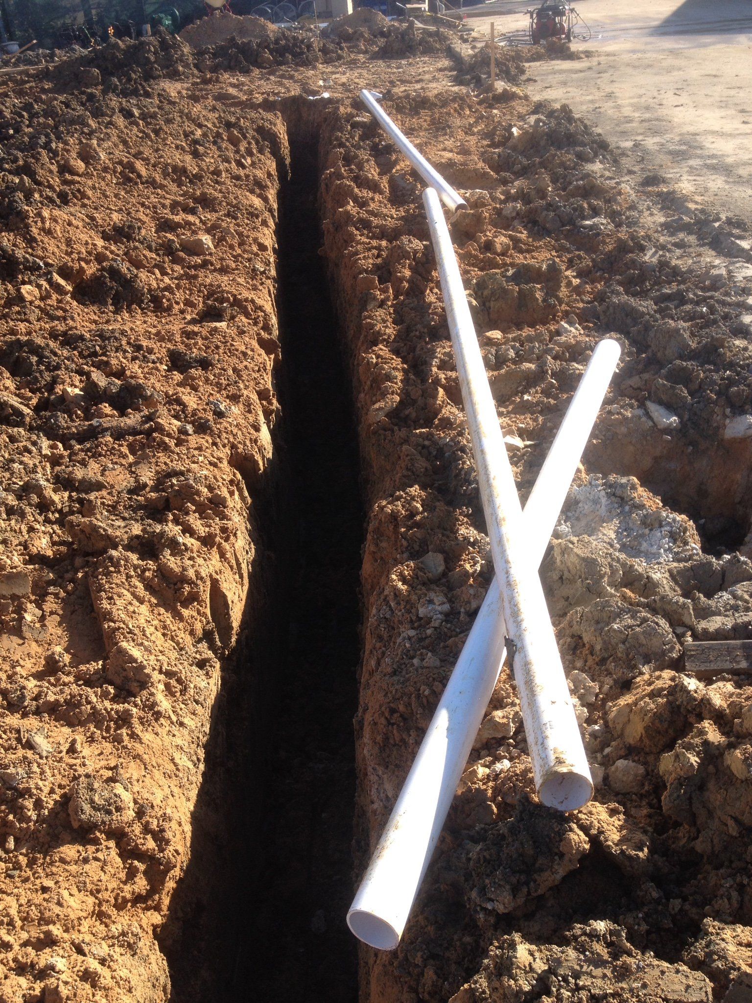 Trenched dirt path with two white PVC pipes laid across the ground outdoors.