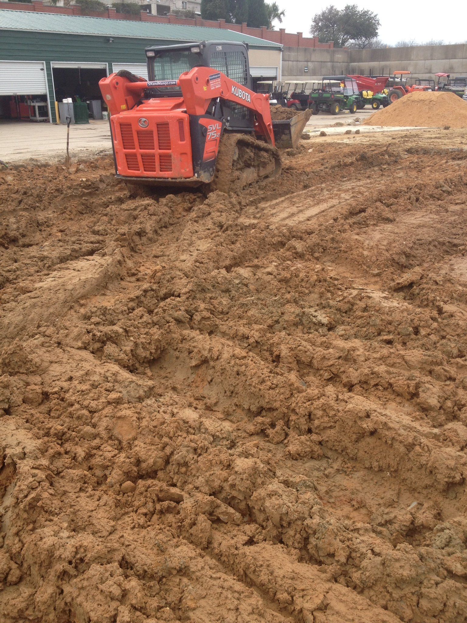 Orange skid steer driving through muddy construction site near buildings and dirt piles