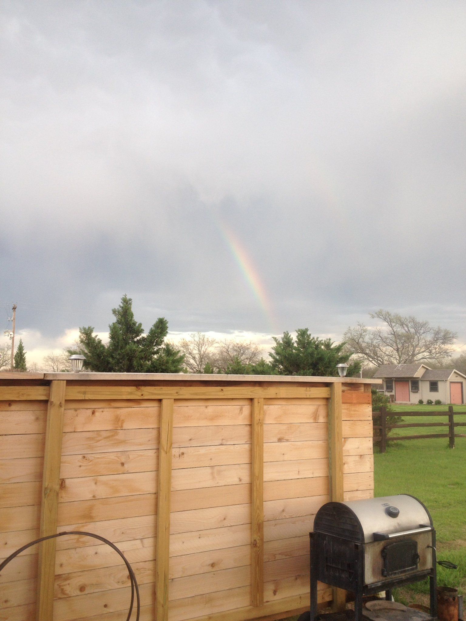 Wooden backyard fence with a grill under cloudy skies and a faint rainbow in the distance
