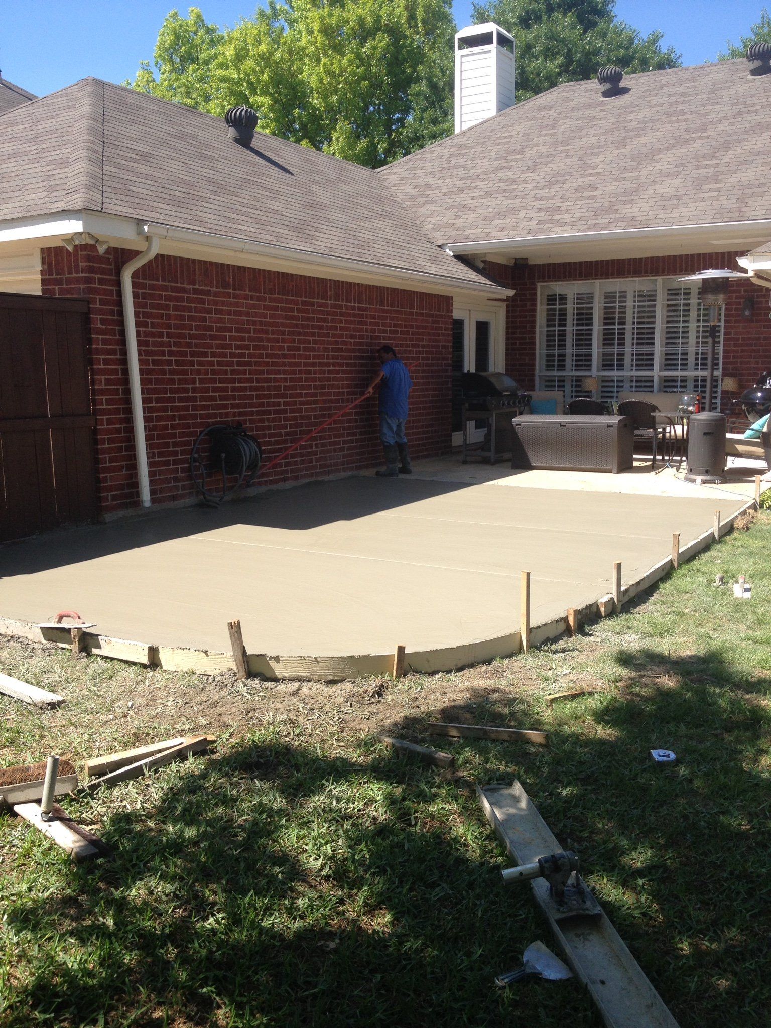 Backyard patio under construction beside a brick house, with a worker near the wall.