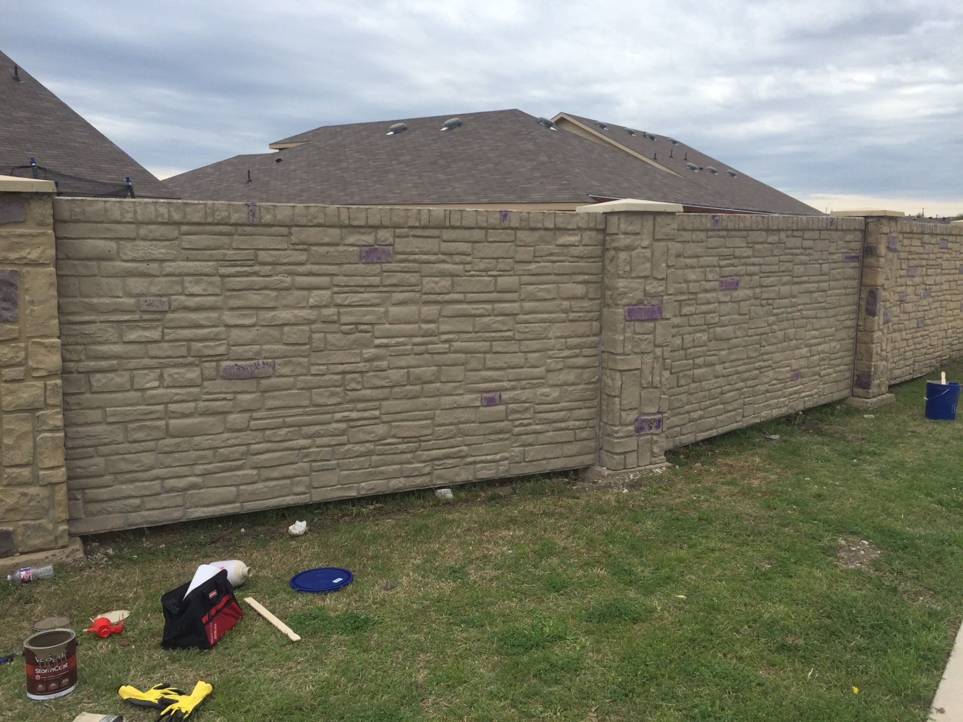 Backyard with tan brick fence and house roof; lawn tools and a blue bucket on the grass.