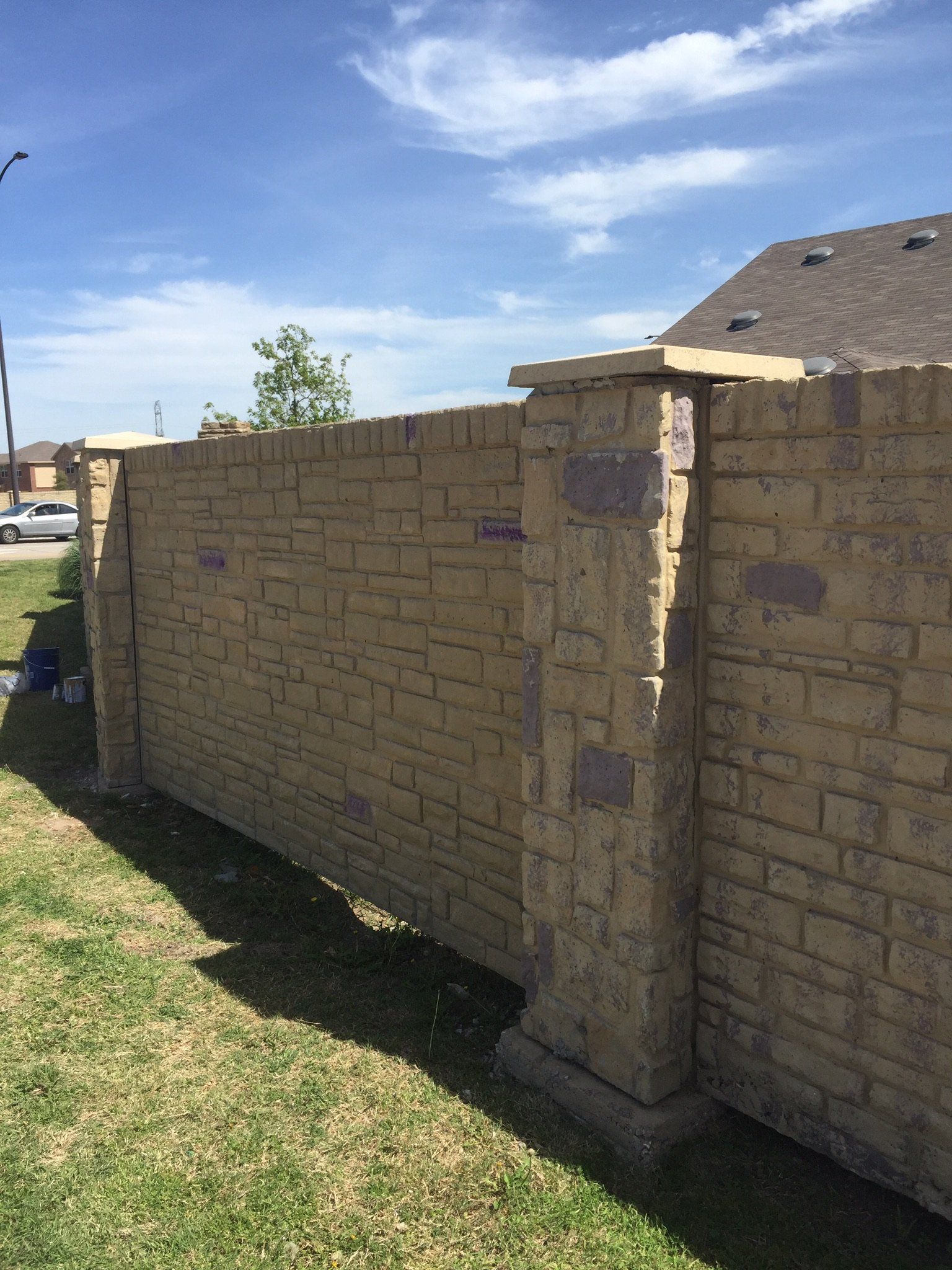Stone privacy wall beside a grassy yard under a blue sky, with a house roof visible behind it