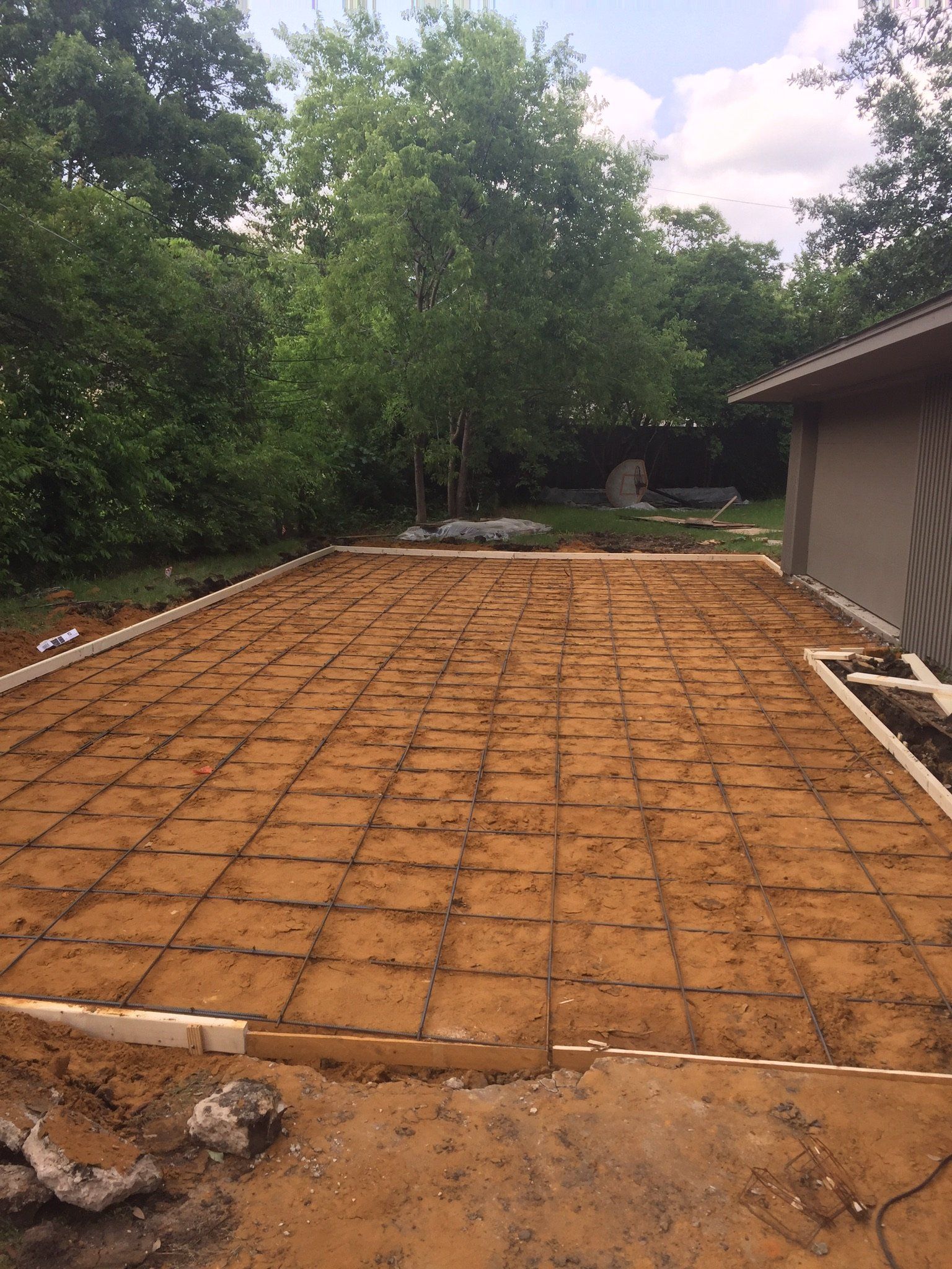 Freshly graded backyard with rebar grid, bordered by dirt, trees, and a house on the right.