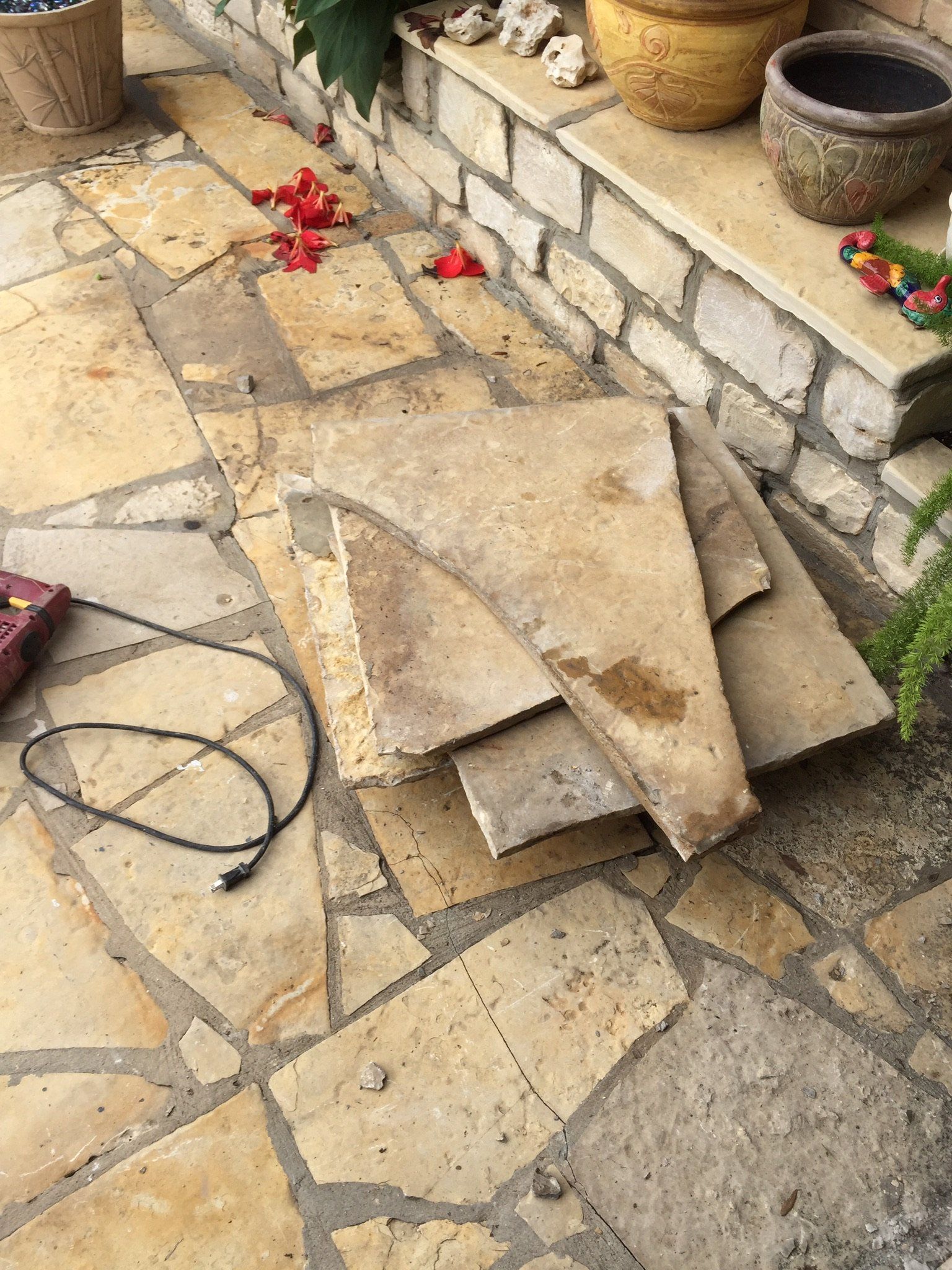 Stacked stone slabs and a black cord on a tiled patio beside a raised planter and potted plants.