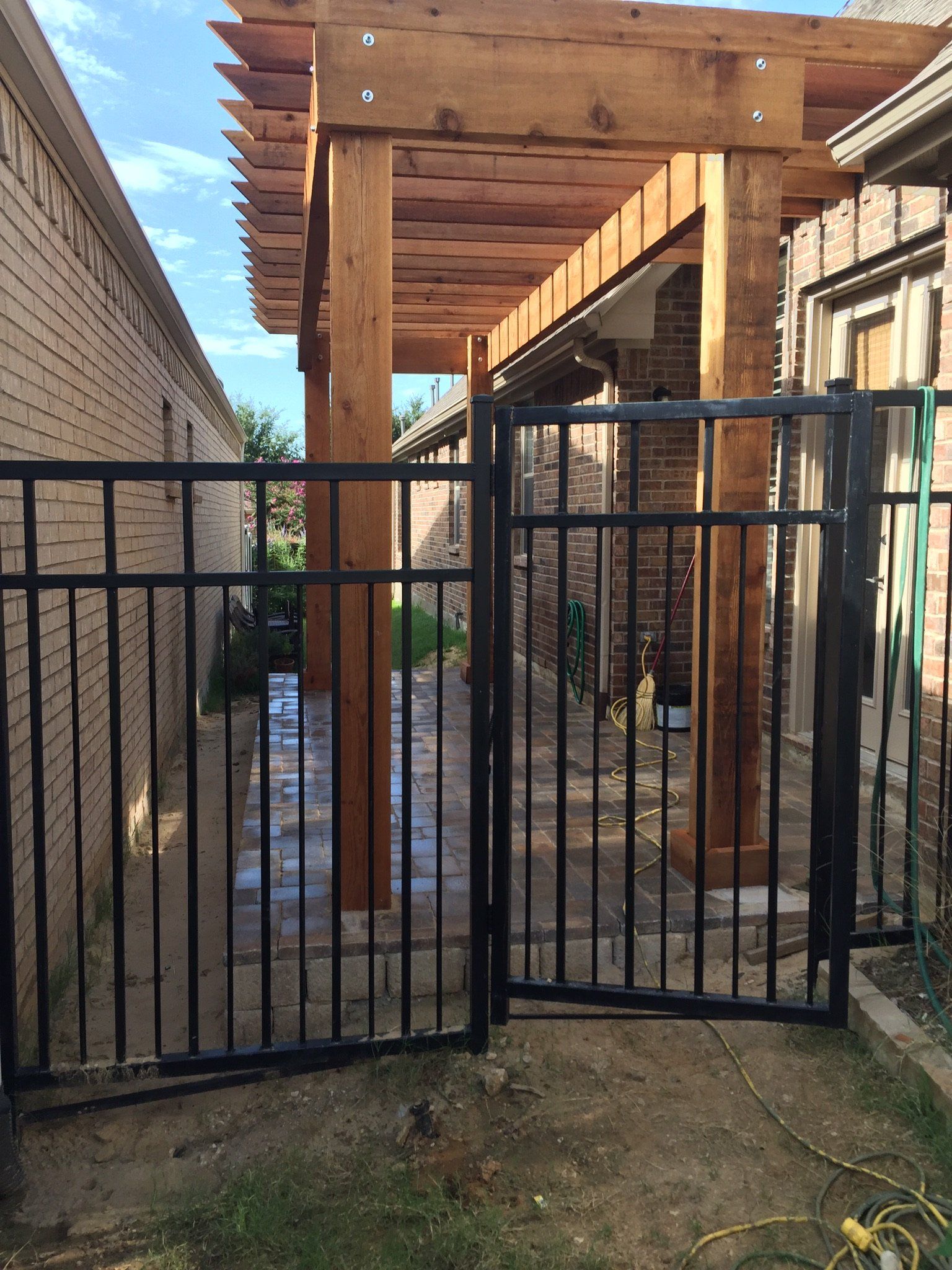 Black metal gate in front of a narrow backyard passage with wooden pergola beams above.
