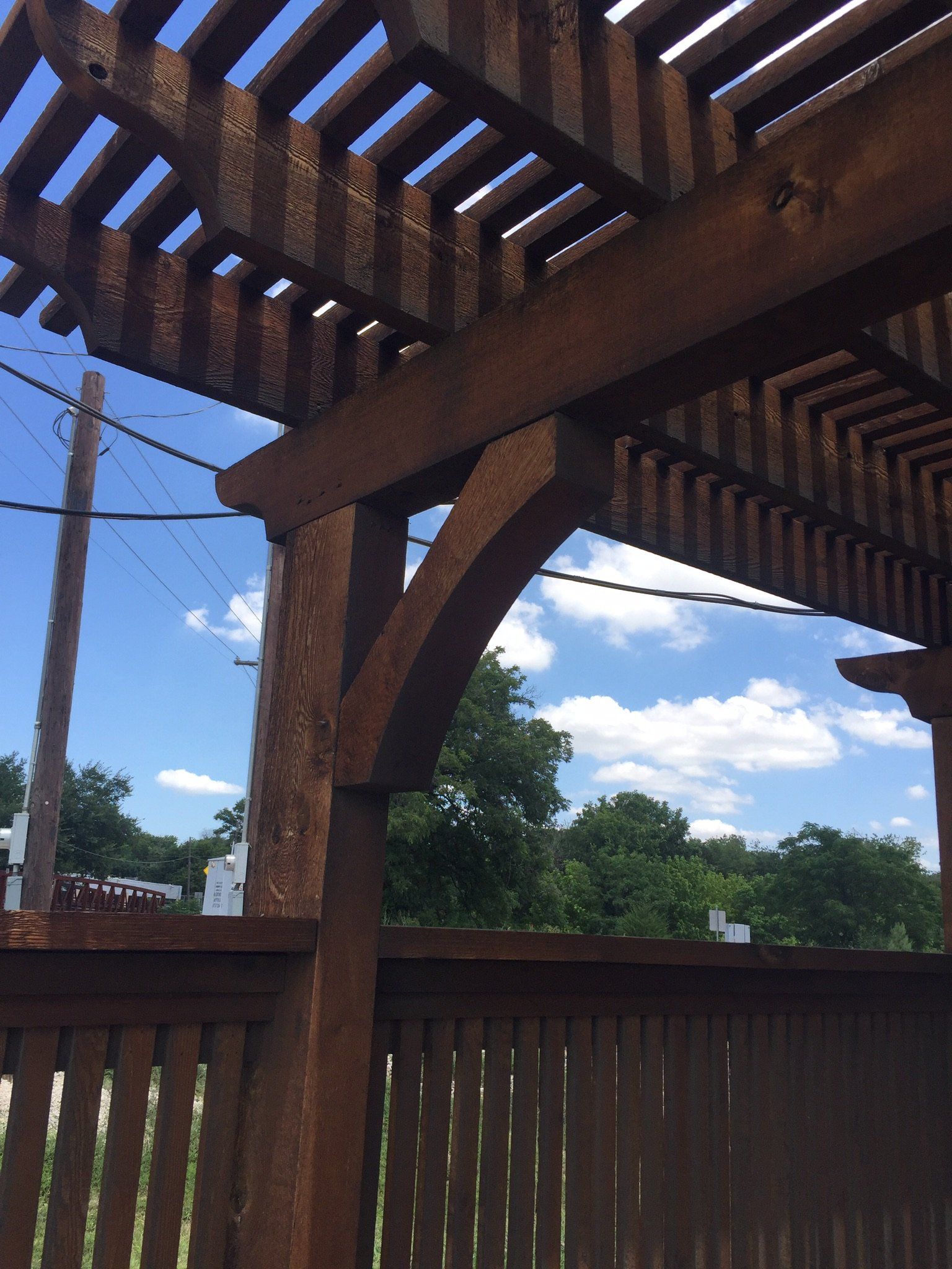 Wooden pergola with slatted roof and fence, overlooking a blue sky with clouds and trees.