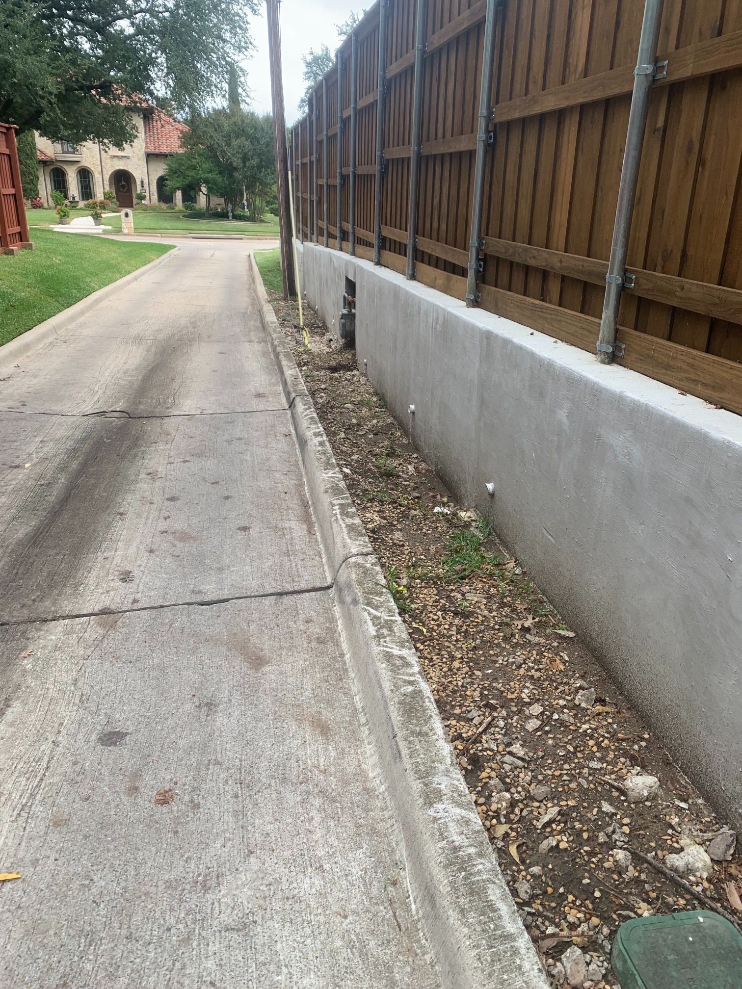 Concrete sidewalk beside a tall wooden fence, with a narrow dirt strip and houses in the background.