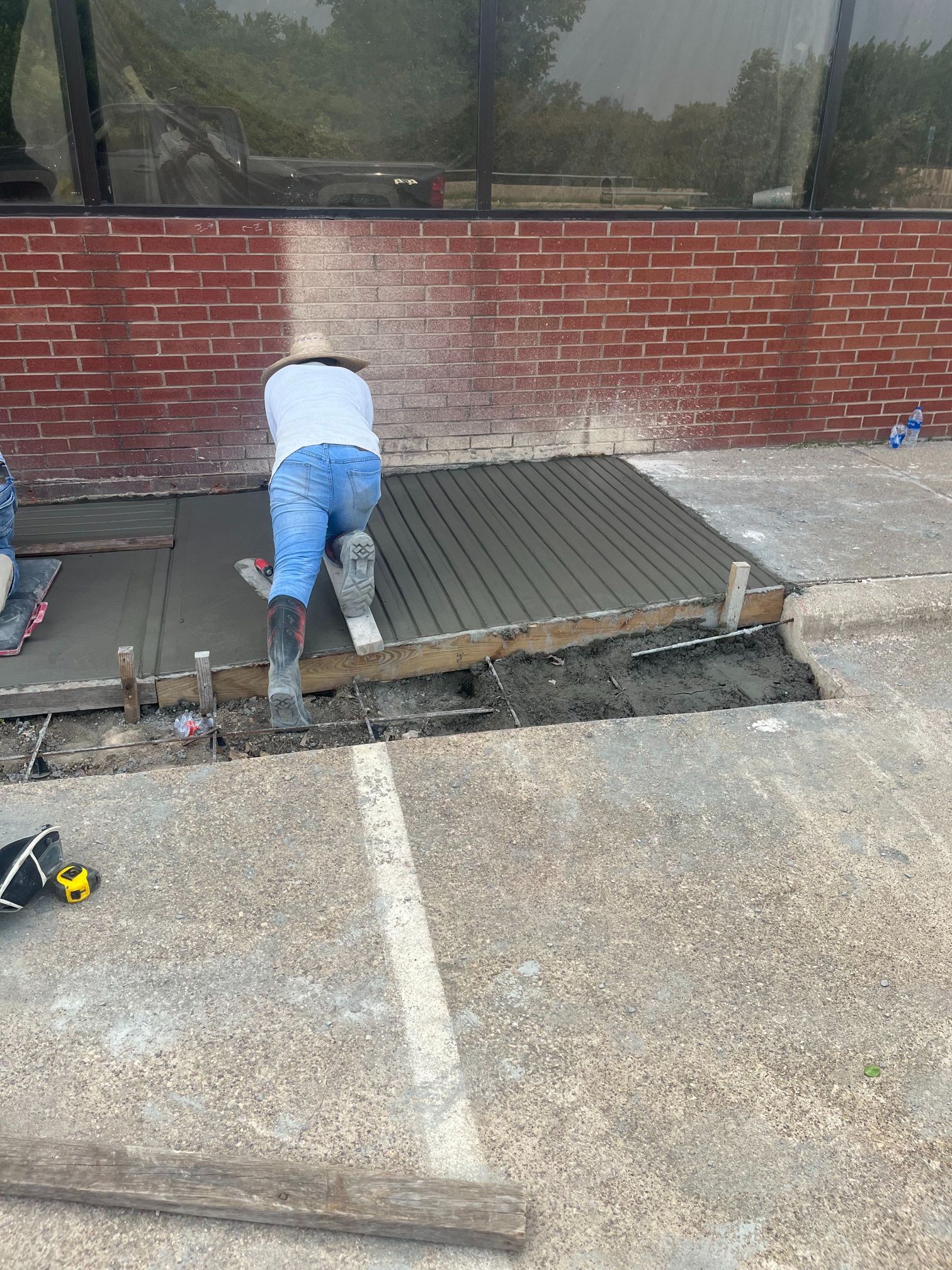 Worker kneeling on a newly poured concrete patio beside a brick building, smoothing the surface.