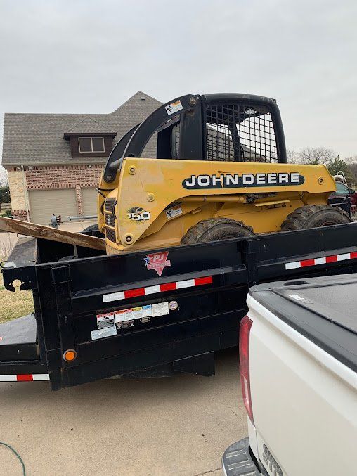 Yellow John Deere skid steer on a black trailer in a driveway beside a white pickup and house.