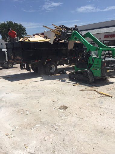 Green skid steer unloading debris into black dump truck in a sunny lot