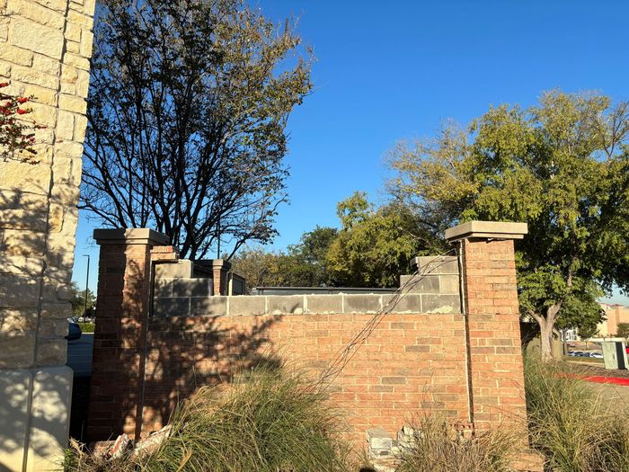 Brick wall and pillars beside a stone building, with trees and blue sky in the background.