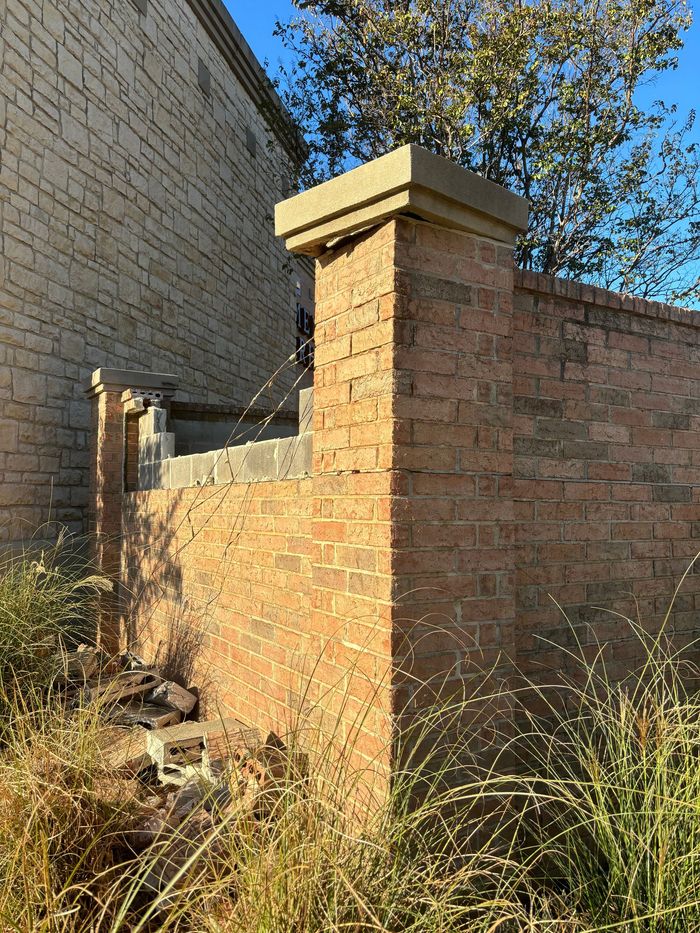 Brick courtyard wall with a square pillar beside a stone building and dry grass in the foreground