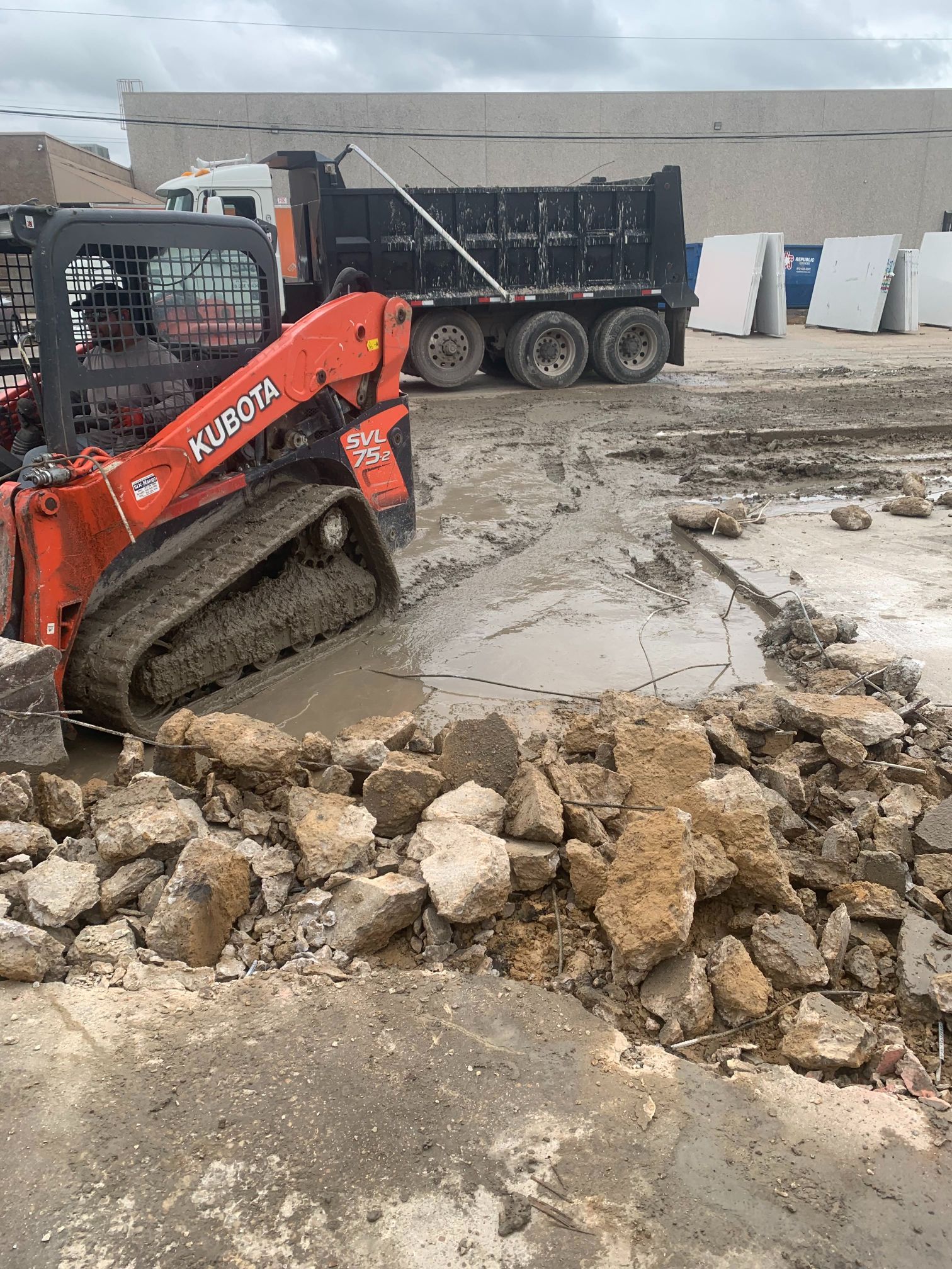 Red skid-steer loader beside a muddy construction site with rubble and a dump truck in the background