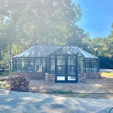 Glass greenhouse with black-framed doors and brick base beside trees on a sunny day