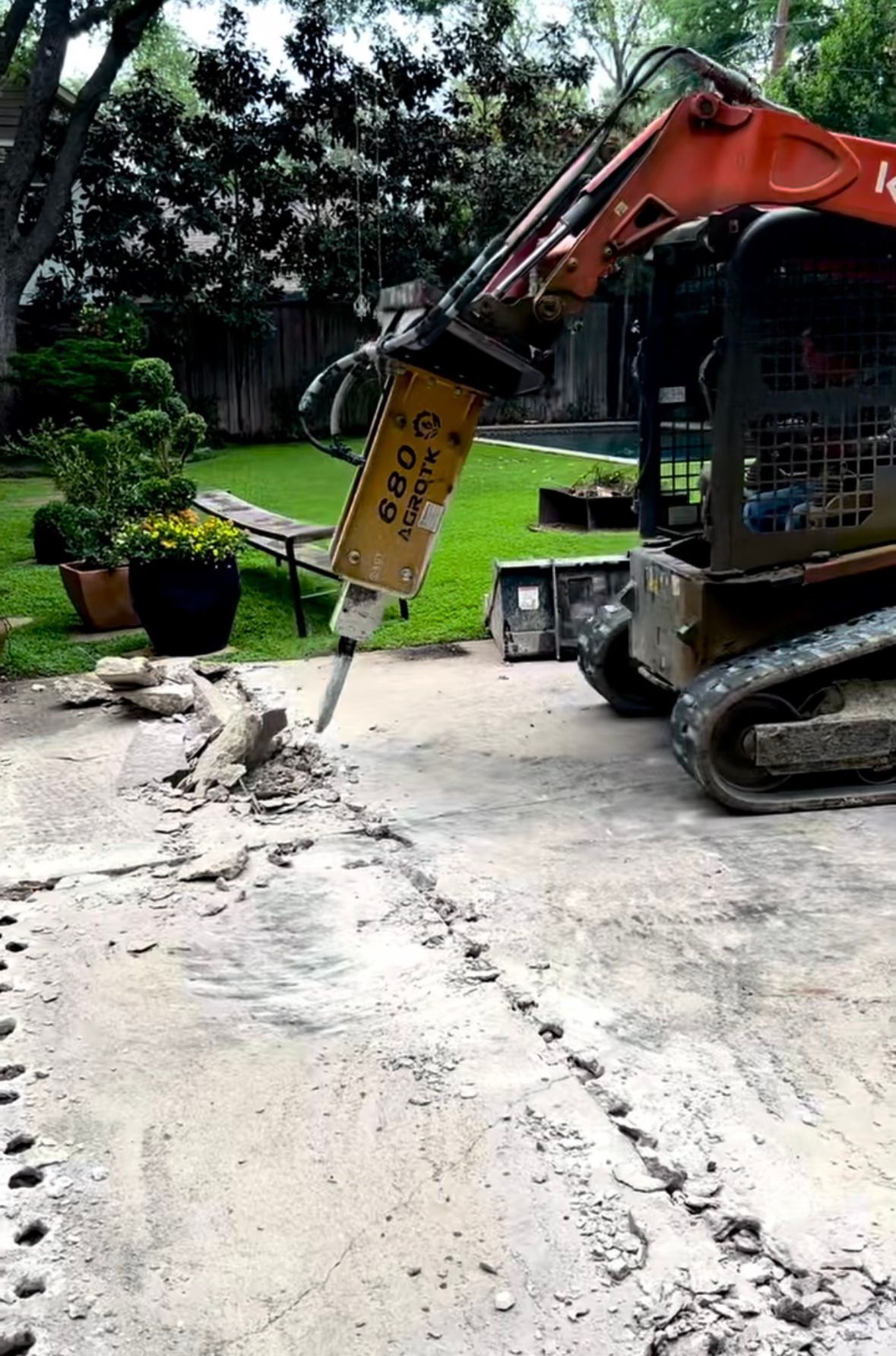 Excavator breaking concrete driveway beside a lawn and potted plants outdoors