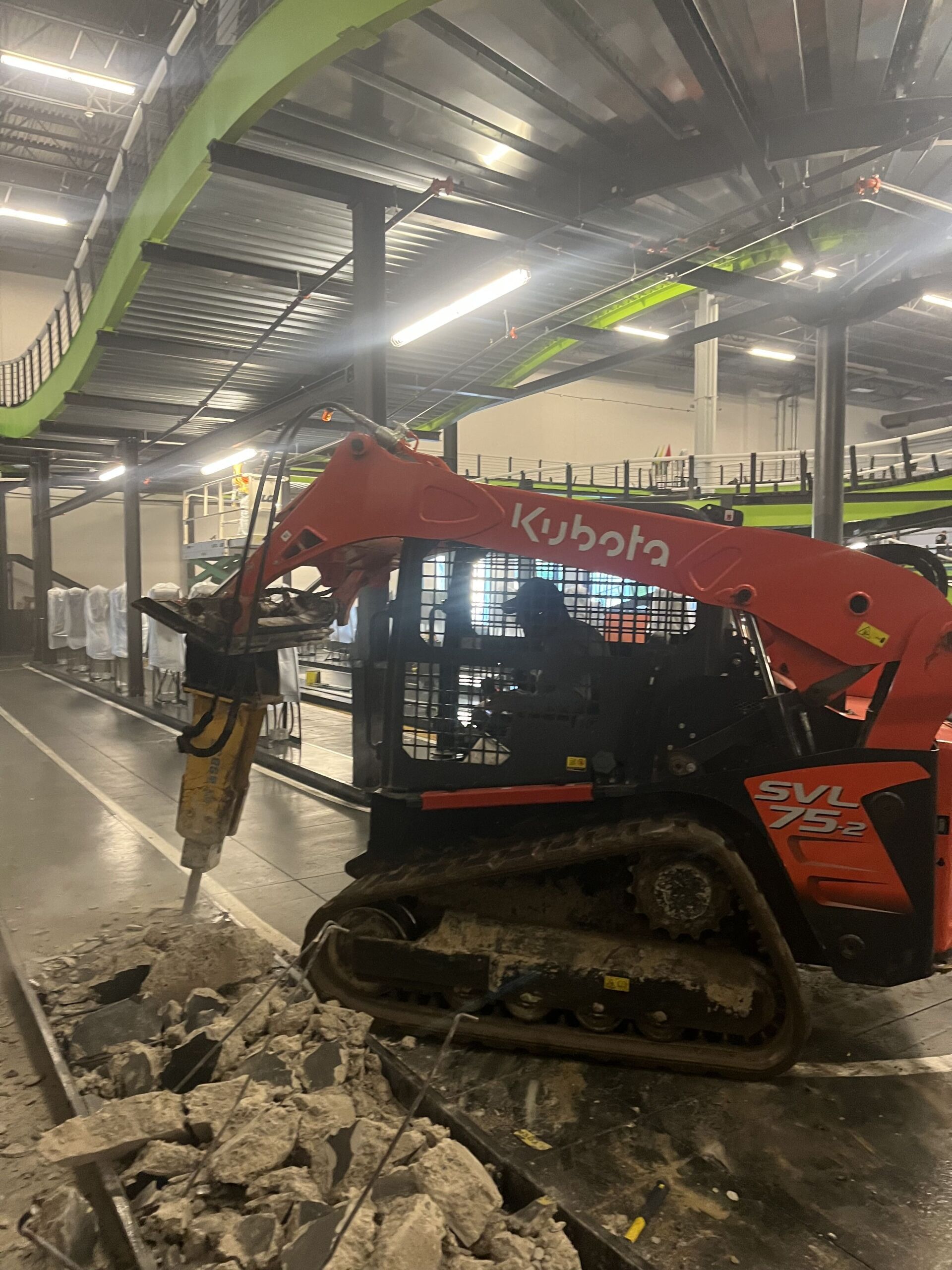Orange Kubota skid steer with jackhammer clearing broken concrete inside a warehouse under construction.