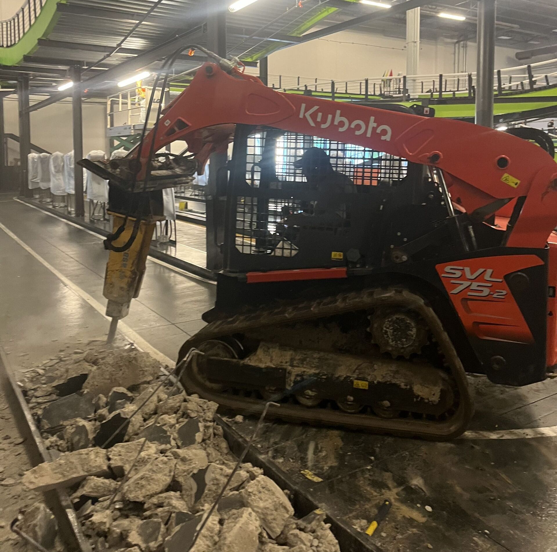 Red Kubota skid-steer breaking concrete inside an industrial building, dust and debris around it.