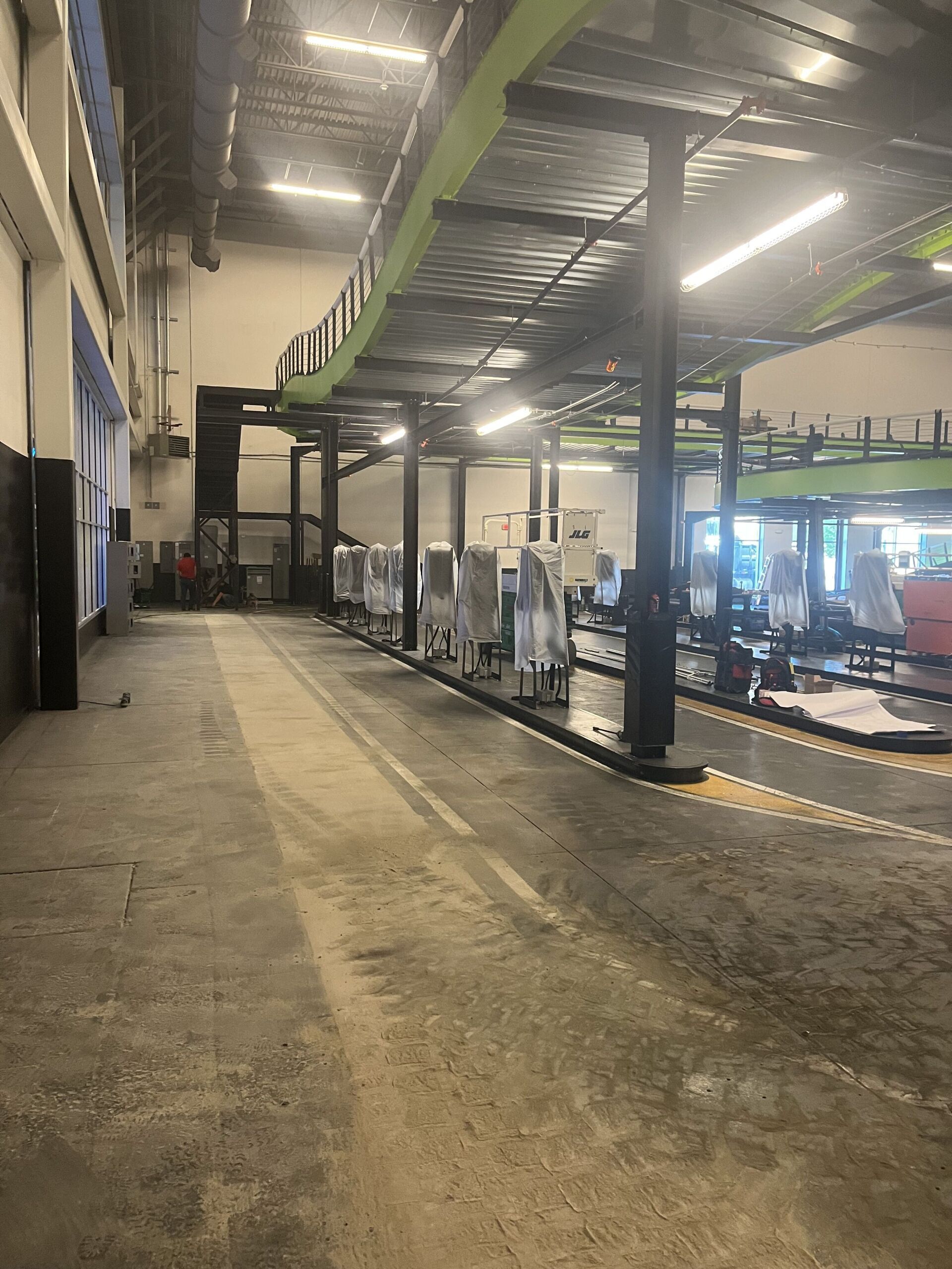 Empty indoor gym floor with equipment lined along the right side and large windows in back.