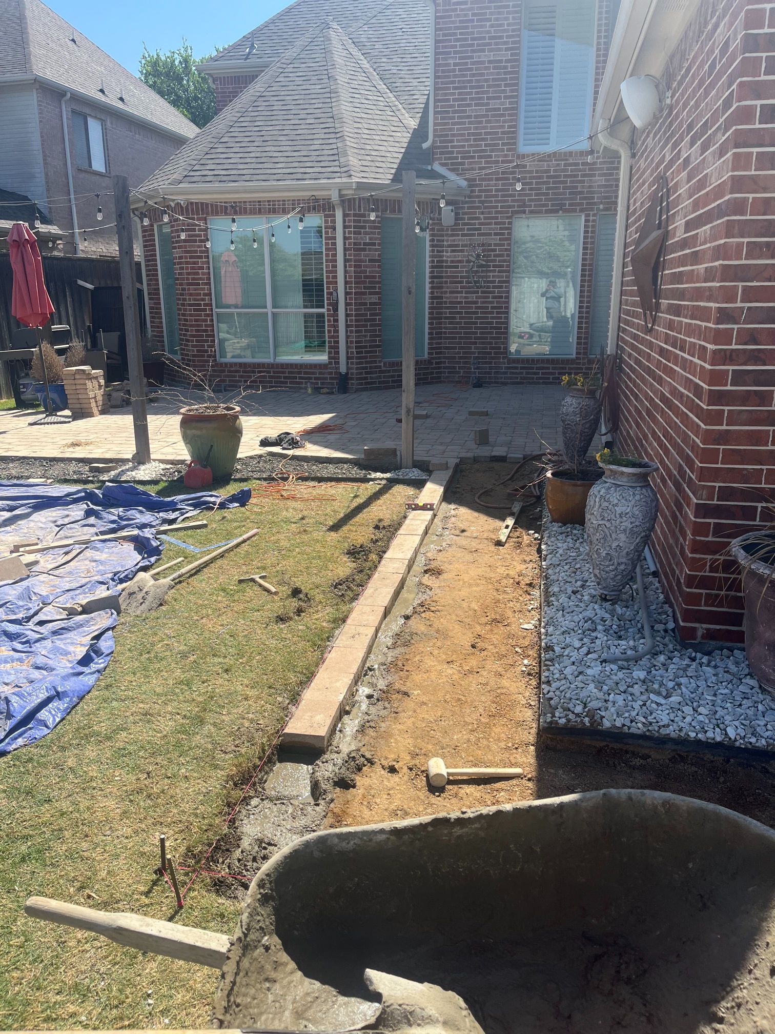 Backyard construction trench beside a brick house, with a tarp, gravel, and exposed soil.