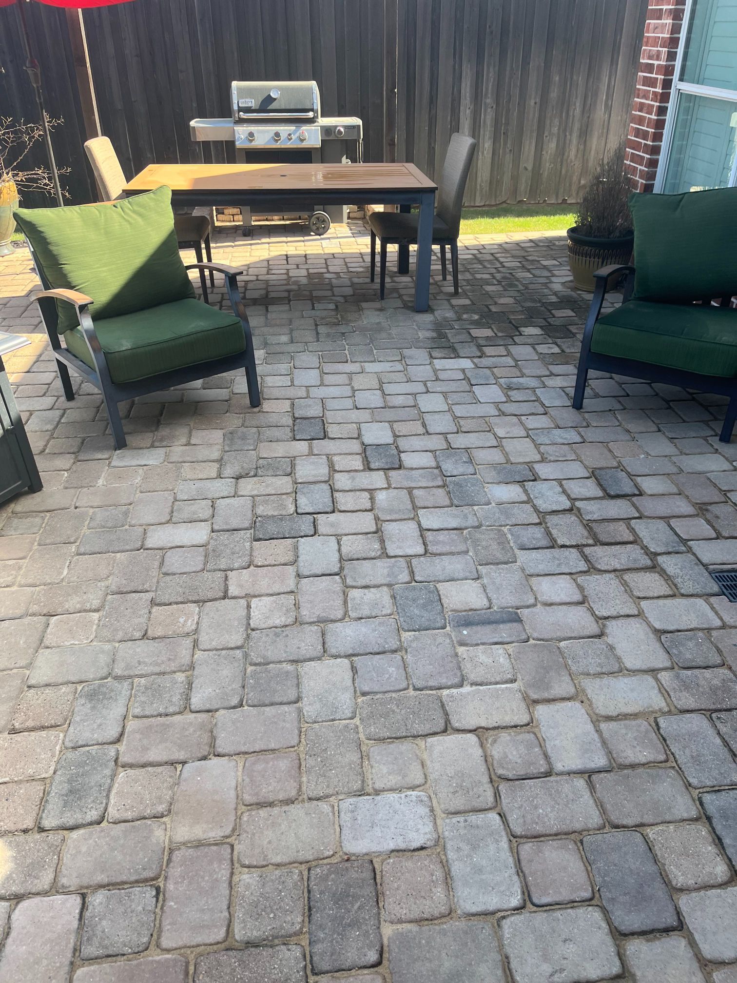 Patio with gray pavers, two green chairs, and a wooden table near a grill under a covered area