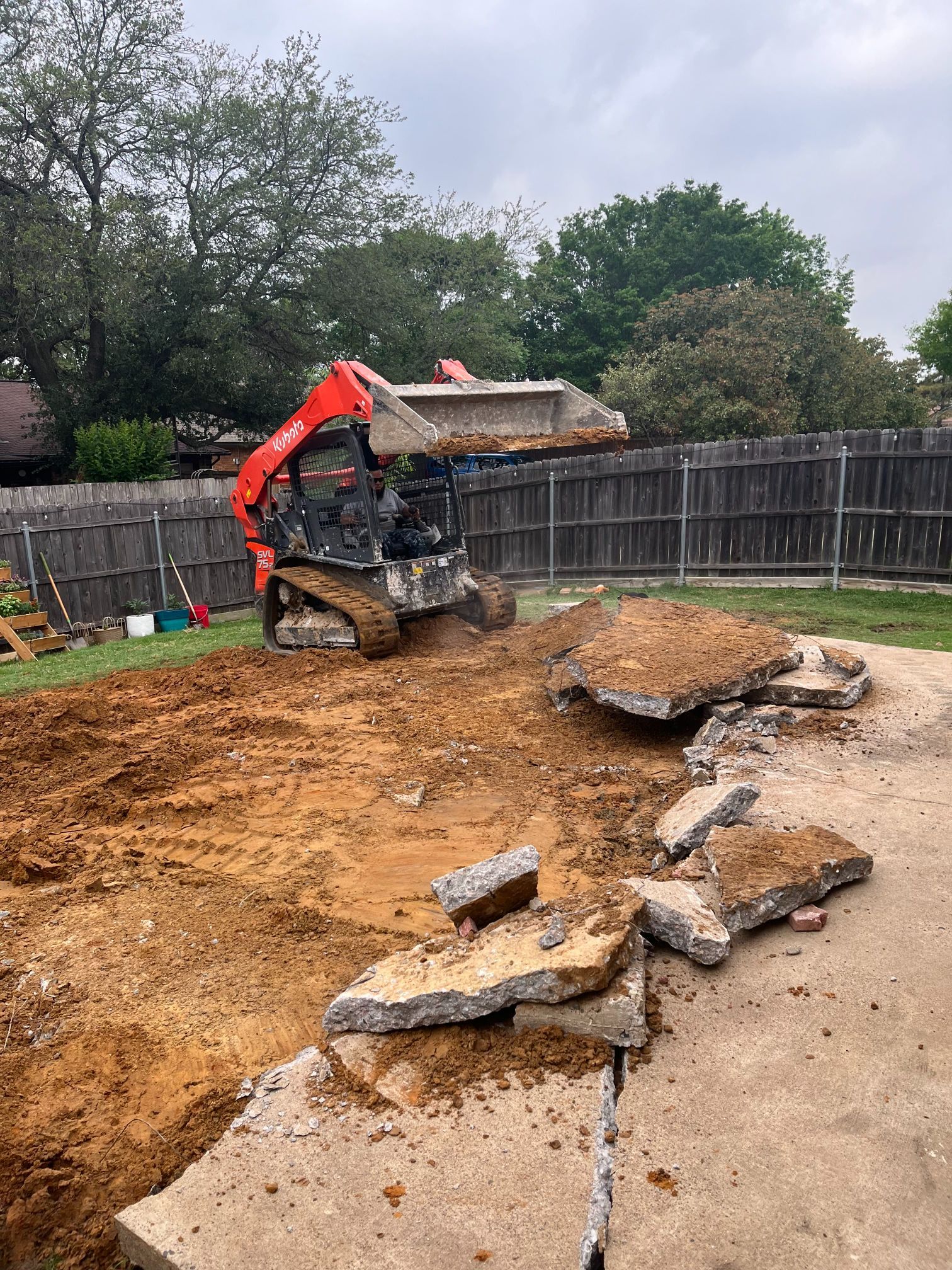 Mini excavator clearing dirt in a fenced backyard beside broken concrete slabs and a driveway