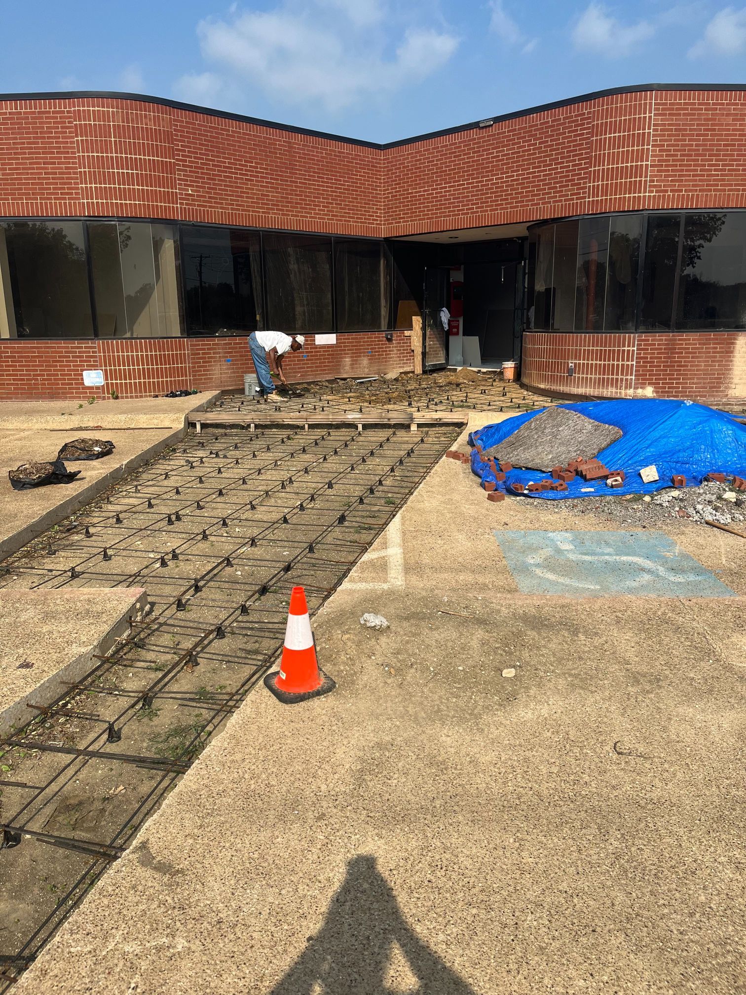 Construction site with rebar grid and orange cone in front of a brick building, covered materials nearby