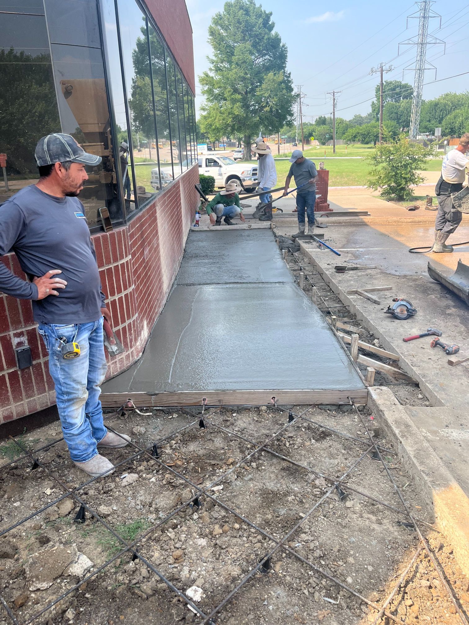 Workers pouring fresh concrete for a sidewalk beside a building, with one man standing nearby.