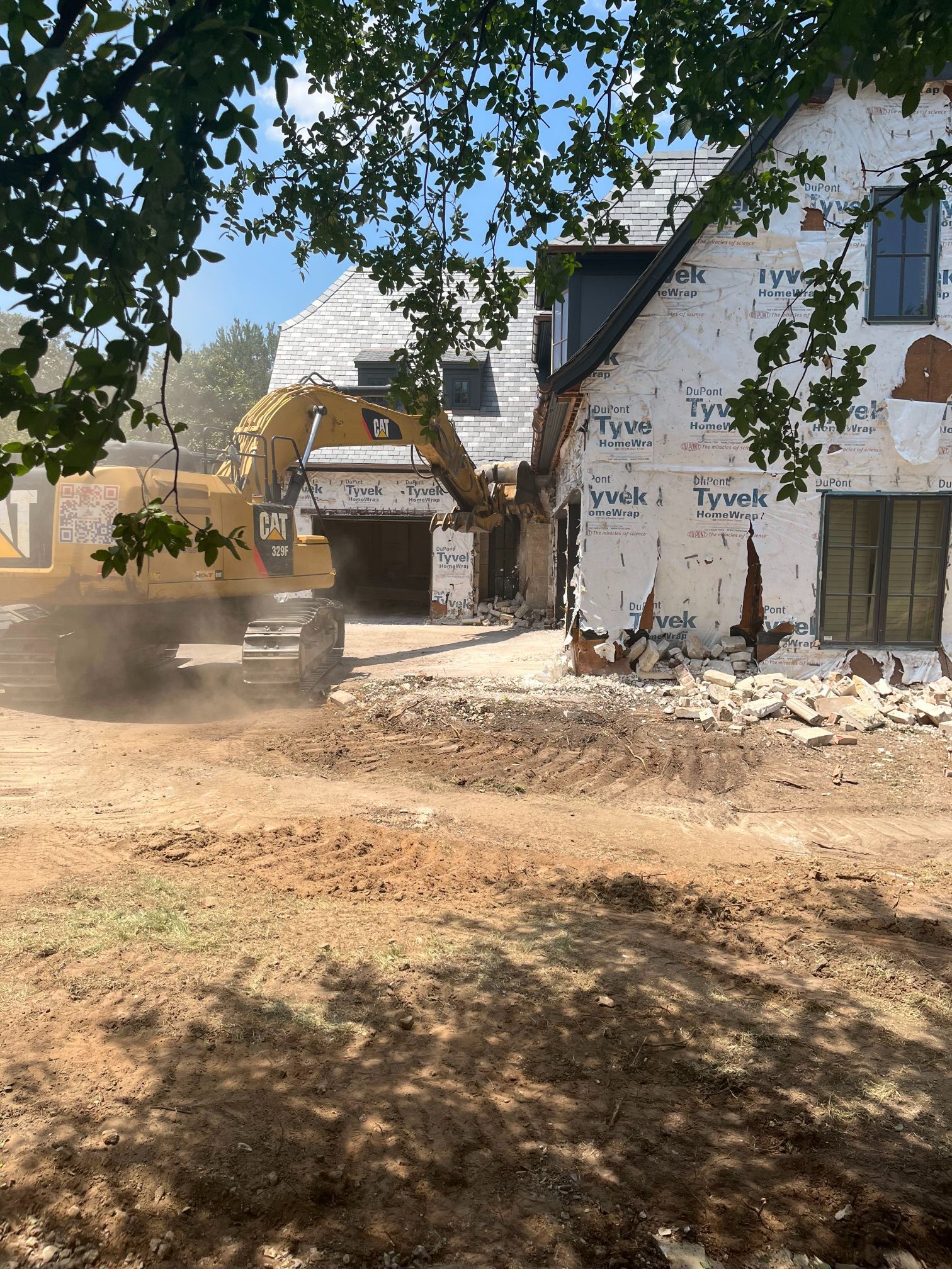 Demolition site with a partially torn-down house, rubble, and dust under a leafy tree canopy